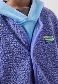 Gros plan sur une personne portant une veste polaire bleue avec un patch "Super Vintage" sur un sweat à capuche bleu clair et une chemise marron en dessous.