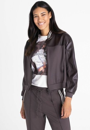 Femme souriante portant une veste en cuir marron foncé par-dessus un t-shirt blanc à motif graphique et un pantalon marron assorti avec des bandes blanches sur les côtés.