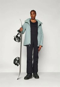 Lys blå skijakke med hette, svarte skibukser og svarte støvler. Holder et snowboard med bindinger, står foran en ensfarget bakgrunn.