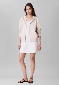 Sweat à capuche beige avec fermeture éclair, deux poches avant et logo sur la poitrine, associé à une robe blanche sans manches et des sandales noires.