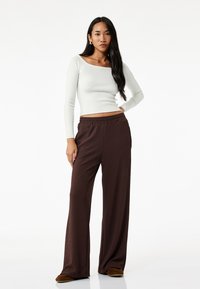Witte geribbelde lange mouw top gecombineerd met bruine wijde broek met een elastische tailleband. De stof heeft een zachte textuur.