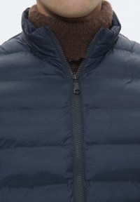 Marineblauer Pufferjacke mit hohem Kragen, ausgestattet mit horizontalen Steppnähten und einem Reißverschluss auf der Vorderseite, getragen über einem braunen, hochgeschlossenen Pullover.