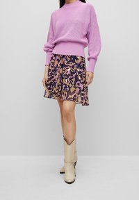 Lila pletený svetr s žebrovanou texturou, doplněný o vzdušnou, vícebarevnou sukni s maskáčovým vzorem a béžové kotníkové boty na podpatku.