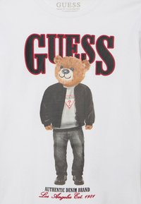 Hvid T-shirt med en bjørn i en sort jakke og grå sweatshirt. Teksten lyder "GUESS" i fede røde og sorte bogstaver med logodetalje.