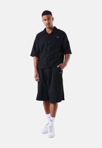 Camisa negra de manga corta con un patrón texturizado y cierre de botones, combinada con pantalones cortos negros. Presenta líneas limpias y acentos de marca mínimos.