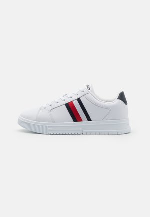 Witte lage sneaker met marineblauwe, rode en witte zijkantenstrepen, witte veters en een zwarte hielstrook op een witte achtergrond.