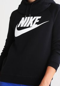 Svart Nike-hoodie med vitt logotyp, bärs av en person med händer delvis synliga och blå byxor.