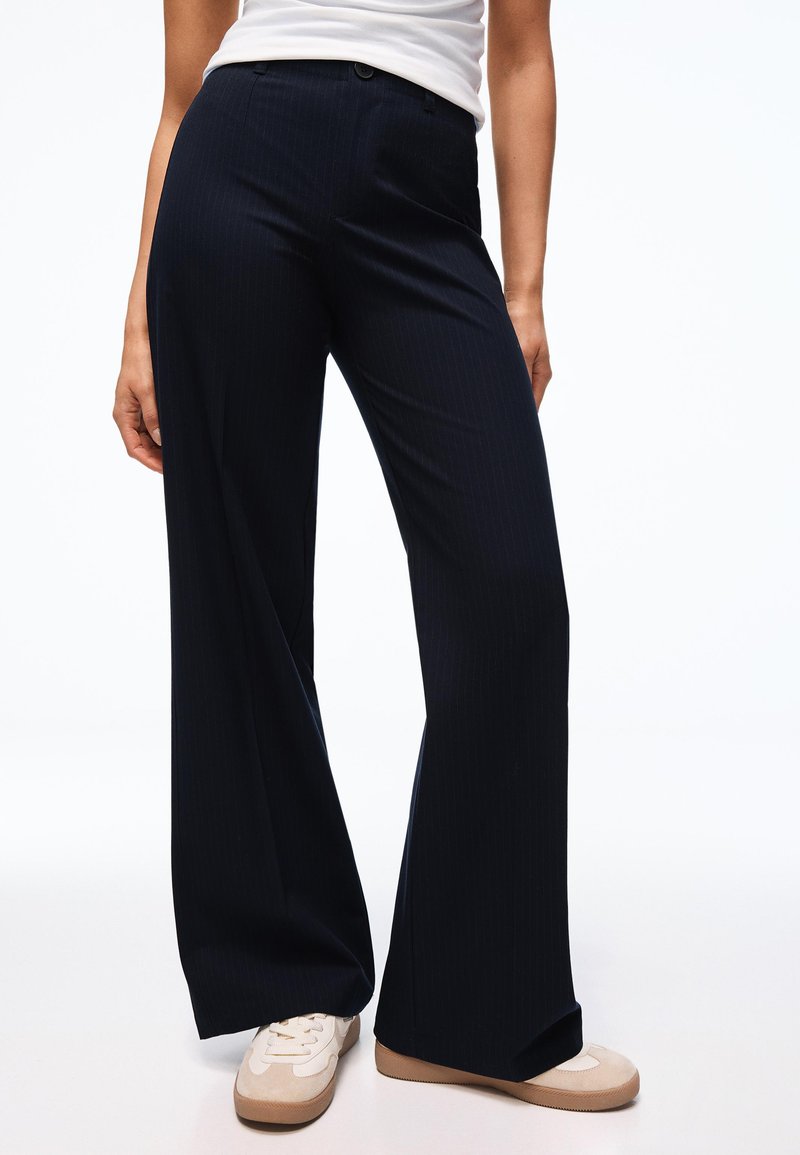 Persoon draagt zwarte wijde broek met hoge taille, gecombineerd met witte sneakers en een wit shirt, staand tegen een effen achtergrond.