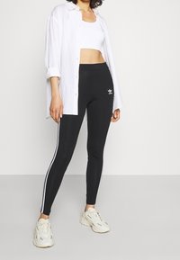 Svarta leggings med vita sidostreck, med en Adidas-logotyp på vänster lår. Matchas med en vit crop top och en lös lång skjorta.