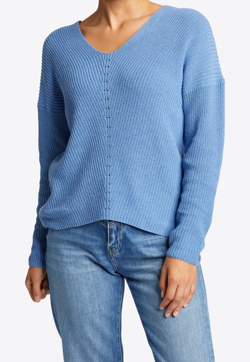 Femme portant un pull en maille bleu clair à col en V avec manches longues et un jean bleu délavé moyen, debout avec une main détendue le long du corps.