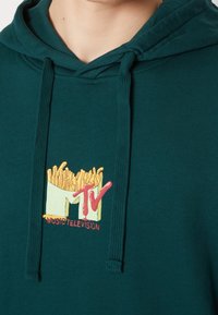 Sweat à capuche vert en coton, avec un motif de frites et le logo "MTV" en rouge et jaune, doté d'un cordon de serrage et de poignets côtelés.