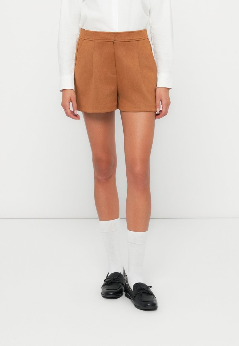 Personne portant un short taille haute beige, une chemise blanche à manches longues, des chaussettes blanches et des mocassins noirs, debout sur un fond blanc.
