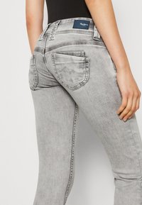 Šedé skinny džíny s vysokým pasem, které mají zadní kapsy a jemné švy, zobrazené ze bočního úhlu.