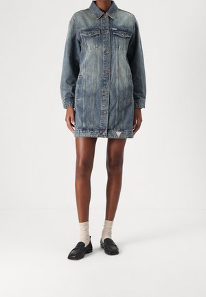 Person trägt ein blaues Jeanskleid mittlerer Länge mit Knöpfen vorne, beige Socken und schwarze Loafers, steht vor einem weißen Hintergrund.