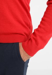 Pull rouge en maille avec ourlet et poignets côtelés, associé à un pantalon marine, avec une main dans la poche, mettant en valeur la texture et le contraste des couleurs.