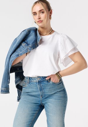 Femme portant un chemisier blanc à manches bouffantes et un jean bleu taille haute, tenant une veste en denim bleue sur l'épaule sur fond blanc.
