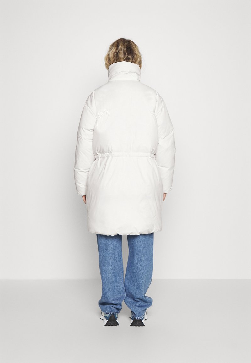 Abrigo puffer blanco con cuello alto, cintura ceñida y tejido texturizado. Combinado con vaqueros azules holgados y zapatos deportivos en blanco y negro.