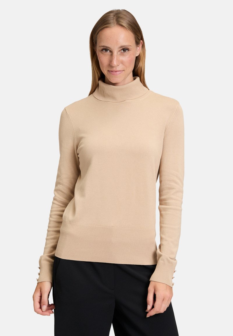 Pull en col roulé beige avec manches longues, texture côtelée, design ajusté et accents de boutons sur les poignets, porté avec un pantalon noir.