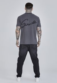 Grijze katoen T-shirt met een zwart cursief logo op de achterkant. Draagt het met donkere jeans en witte sneakers. Tatoeages zichtbaar op de armen.