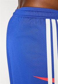 Blauwe sportieve shorts van mesh stof met een gestructureerd oppervlak, witte strepen aan de zijkanten en een rode logo-accent.