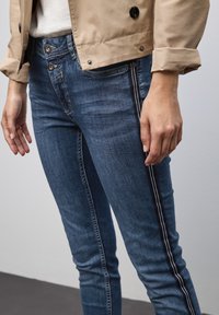 Vaqueros de mezclilla azul con corte slim, con una franja lateral en blanco y negro y cierres de botón de metal en la cintura. Textura suave y acabado liso.