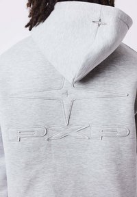 Vue de dos d'une personne portant un sweat à capuche gris clair avec des lettres PXRD en relief et un motif brodé en forme d'étoile sur le dos et la capuche.