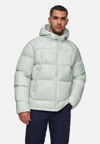 Man in een lichtgrijze gewatteerde hooded jack met ritszakken en donkere broek, staande voor een witte achtergrond.