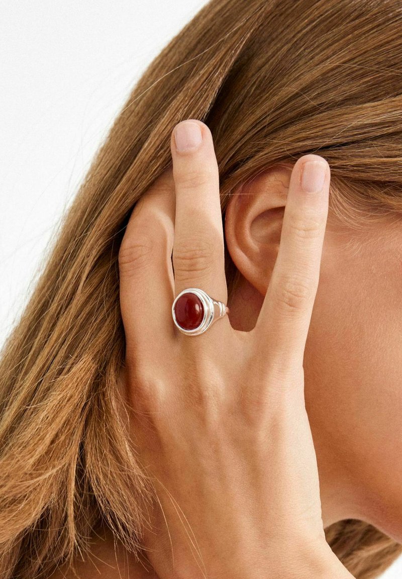 Bague en argent ornée d'une pierre rouge ronde, avec un design en couches, portée à un doigt à côté d'une chevelure brune ondulée et près d'une oreille.