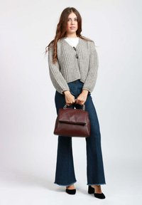 Cardigan corto in maglia grigia indossato sopra una camicia bianca, abbinato a jeans scuri a zampa. Tenendo in mano una borsa a tracolla in pelle bordeaux con manico a catena argento.