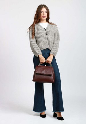 Cardigan corto in maglia grigia indossato sopra una camicia bianca, abbinato a jeans scuri a zampa. Tenendo in mano una borsa a tracolla in pelle bordeaux con manico a catena argento.