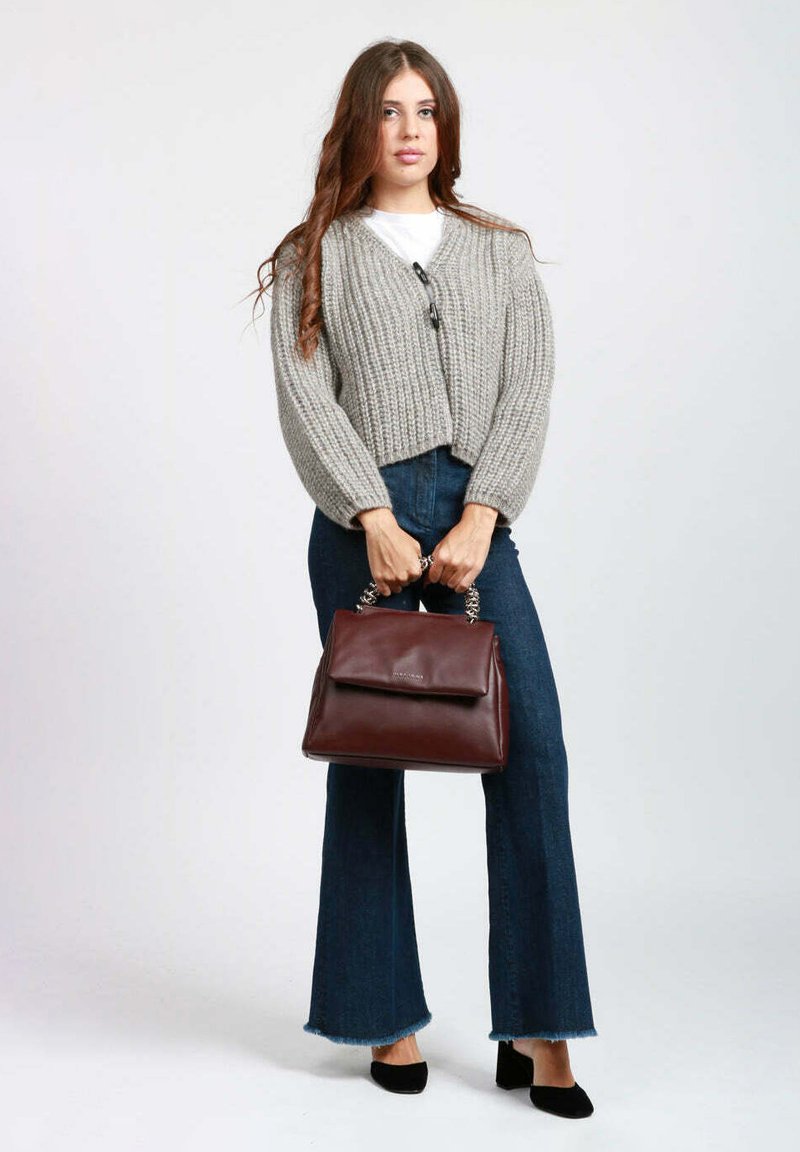 Cardigan corto in maglia grigia indossato sopra una camicia bianca, abbinato a jeans scuri a zampa. Tenendo in mano una borsa a tracolla in pelle bordeaux con manico a catena argento.