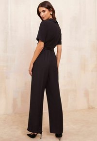 Tuta nera con maniche corte, colletto, gambe larghe e vita accentuata da una cintura. Texture liscia con una vestibilità sartoriale. Indossata con ballerine a punta.