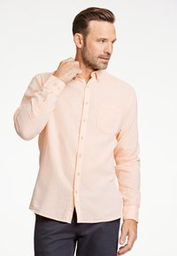 Camisa de botones color melocotón claro hecha de tela texturizada, con un bolsillo en el pecho y mangas largas, combinada con pantalones oscuros.