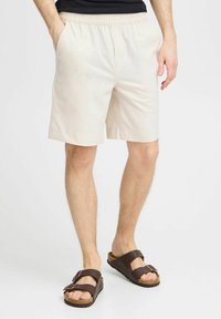 Pantalones cortos de algodón beige con una cintura elástica, bolsillos laterales y un corte relajado, combinados con sandalias marrones con hebilla y suela de corcho.