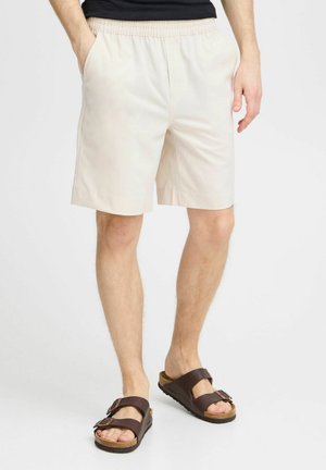 Shorts din bumbac bej cu talie elastică, buzunare laterale și o croială lejeră, purtate cu sandale maro cu cataramă, prezentând o talpă din plută.