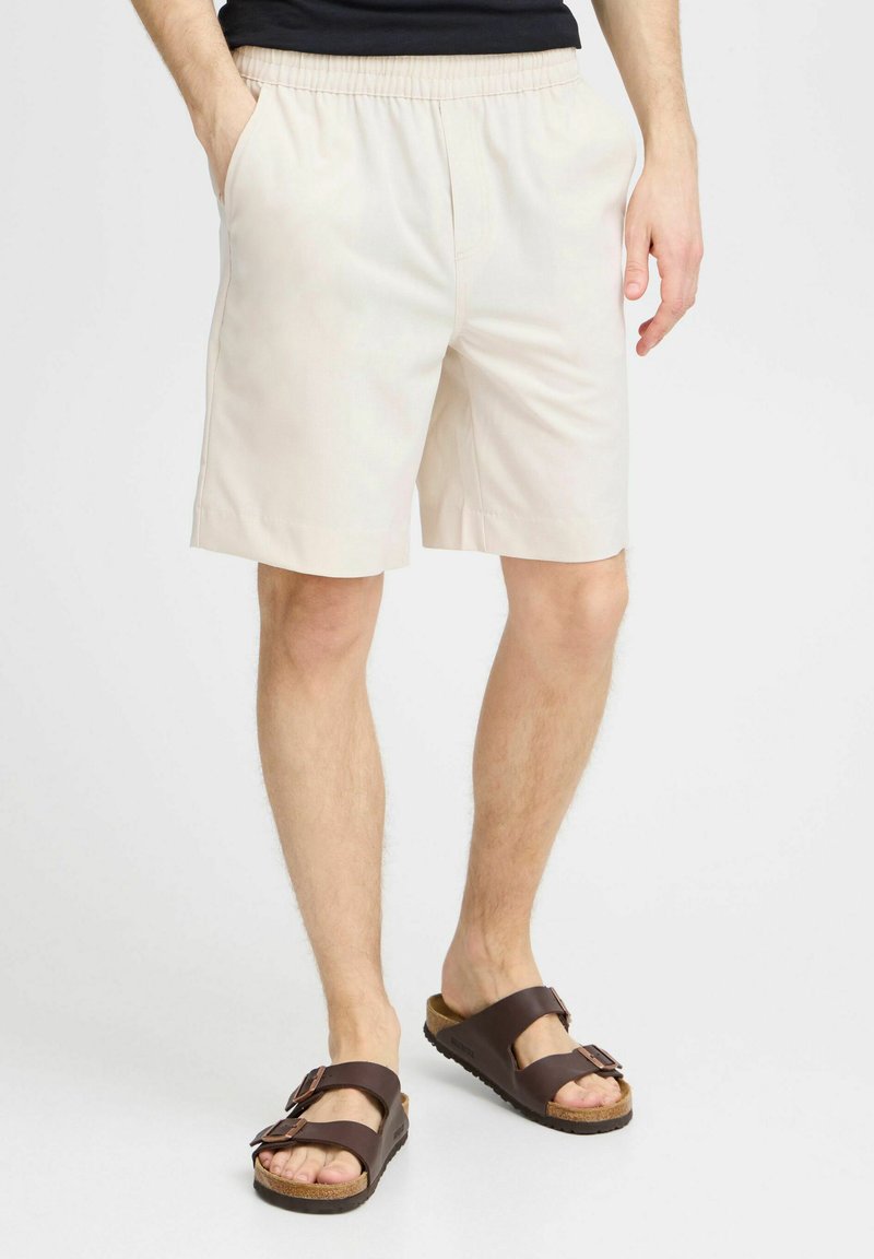 Pantalones cortos de algodón beige con una cintura elástica, bolsillos laterales y un corte relajado, combinados con sandalias marrones con hebilla y suela de corcho.