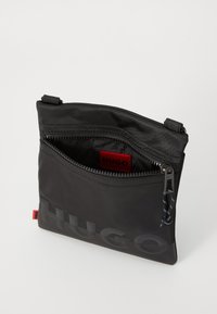 Poche en nylon noir avec fermeture éclair, comportant une étiquette de logo rouge et une texture lisse. Contient un intérieur visible avec un imprimé de logo.