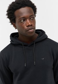 Młody czarnoskóry mężczyzna z kręconymi włosami, noszący czarną bluzę z kapturem z małym haftowanym logo konia, patrzący lekko w lewo.