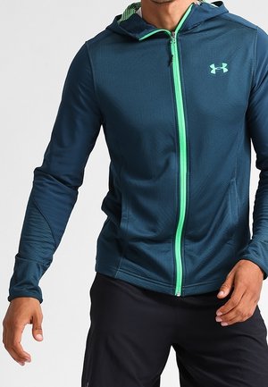 Homme portant une veste à capuche bleu foncé avec une fermeture éclair vert vif et un logo, associée à un pantalon noir, debout, les mains détendues.