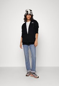 Sudadera negra con cremallera, camiseta blanca, jeans azul claro y zapatillas beige. Sombrero de cubo con patrón en blanco y negro. Atuendo casual con estilo atlético.