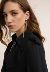 Czarny płaszcz typu trencz z klapowym kołnierzem i ozdobnymi guzikami, zestawiony z długimi srebrnymi kolczykami z frędzlami i kulistym akcentem.