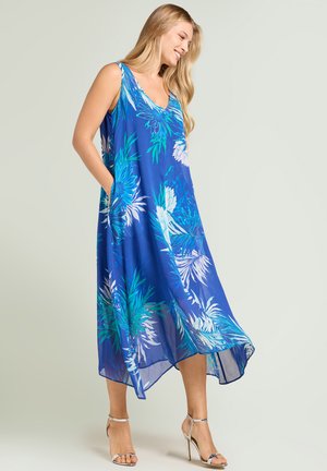 Vrouw in blauwe mouwloze bloemenjurk met zakken, zilveren hoge hak sandalen dragend, glimlachend en naar beneden opzij kijkend.