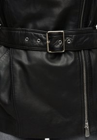 Μαύρο δερμάτινο σακάκι με ραμμένη ζώνη που διαθέτει μεταλλική αγκράφα και πολλαπλές οπές, μαζί με ένα ασημί φερμουάρ στη δεξιά πλευρά.