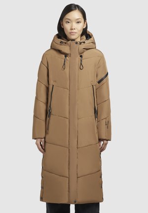 Jeune femme portant un long manteau d'hiver marron rembourré avec capuche, poches zippées et boutons pression sur les côtés, se tenant devant un fond uni.