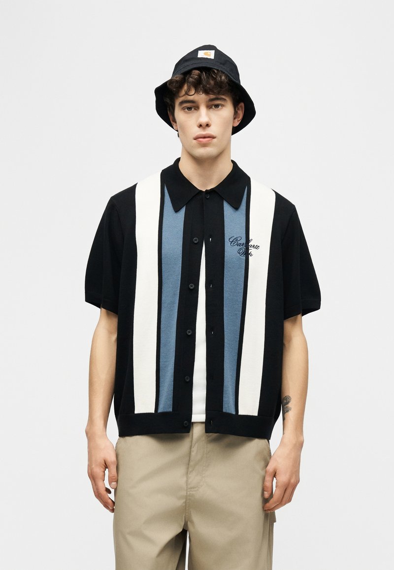 Ung mand iført en sort bucket-hat, sort kortærmet skjorte med hvide og blå lodrette striber og beige bukser, stående.