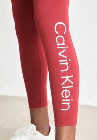 Benen in rode Calvin Klein leggings met witte logo-tekst en witte sneakers op een lichte vloer en wand achtergrond.