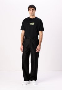 Svart t-skjorte med grønn grafisk tekst, kombinert med svarte vide bukser. Modellen har på seg hvite joggesko og står mot en ensfarget bakgrunn.
