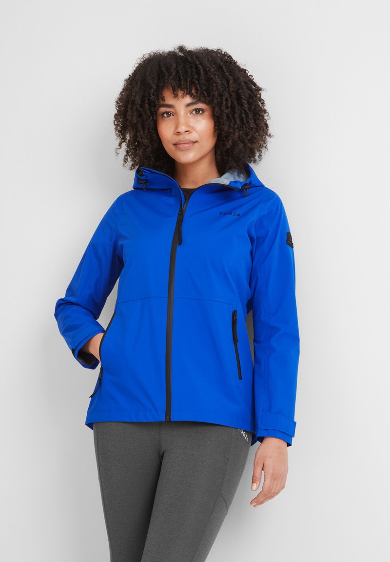 Woman with curly hair wearing a bright blue hooded jacket and dark gray leggings, standing with one hand in a pocket against a white background.