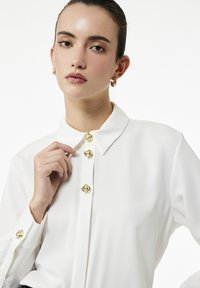 Valkoinen paita, jossa on klassinen kaulus, kullanvärisillä nappiaksentilla ja hihansuu yksityiskohdilla, valmistettu sileästä kankaasta. Yksinkertainen, elegantti muotoilu.