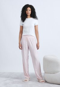 Λευκή υφασμάτινη μπλούζα με κοντά μανίκια, σε συνδυασμό με ροζ ριγέ πυτζάμες. Το μοντέλο φοράει ανοιχτά παντοφλάκια. Απαλό χαλί και στρογγυλή καρέκλα στο φόντο.